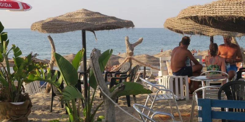 People sitting under straw umbrellas, relaxing on a sandy beach with green plants and ocean view at Camping & Residence Villa Teti