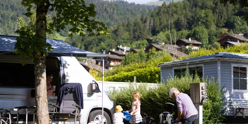 Family interacting beside a camper van, set amidst lush greenery and mountains on a sunny day at Clos du Pin