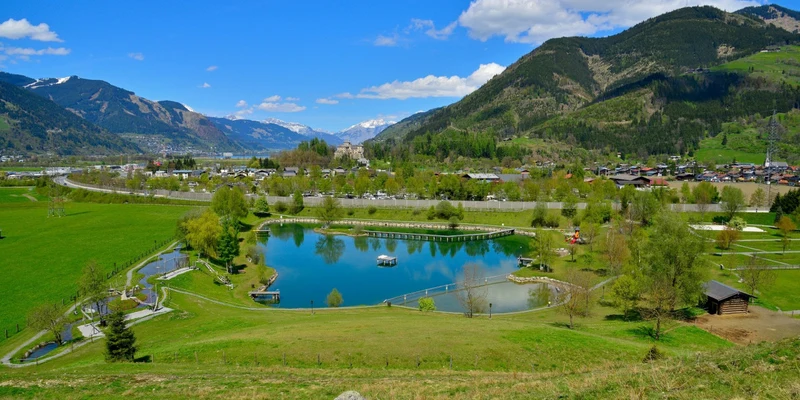 A tranquil pond reflecting mountains and trees, surrounded by green fields and a small village, at Sportcamp Woferlgut