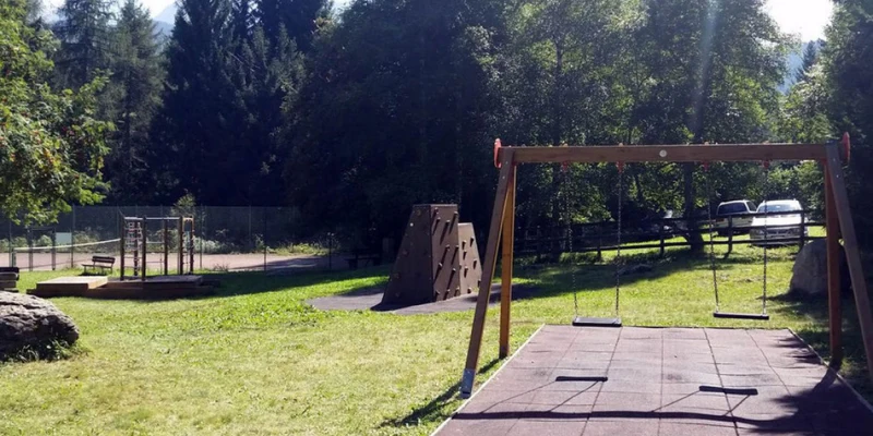Swings stand still on a grassy playground with a climbing wall and benches at Fiemme Village