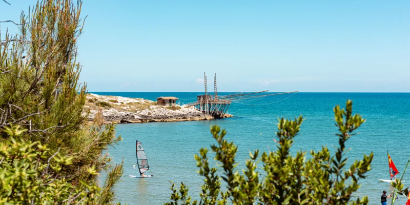 Windsurfer glides across the blue sea near a rocky shore with a wooden structure; trees frame the scene at Camping Village Punta Lunga