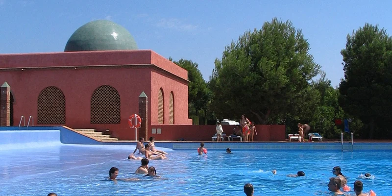 People swimming in a large blue pool by a red building with a green dome, surrounded by trees at El Templo del Sol