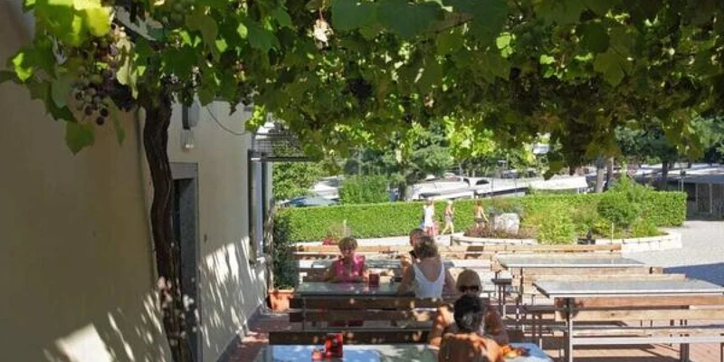 People sitting at picnic tables under a vine-covered pergola, enjoying a meal at Lido Verbano