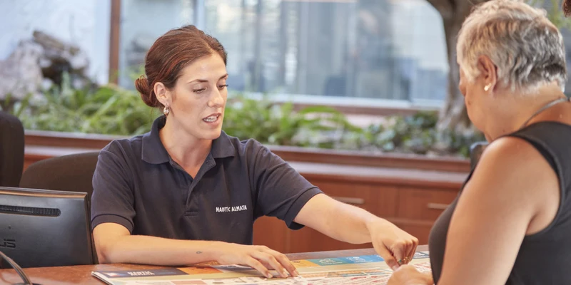 A woman in a uniform points at a map, assisting a woman in a sleeveless top at a wooden counter at Nautic Almata