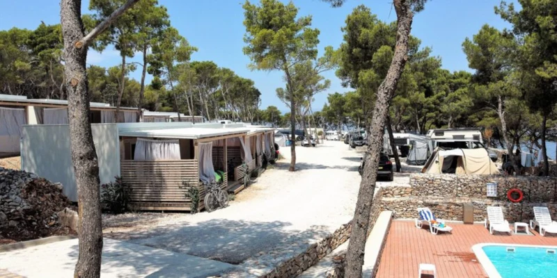 Bungalows and tents line a tree-shaded path near a swimming pool with lounge chairs at Boutique Camping Bunja