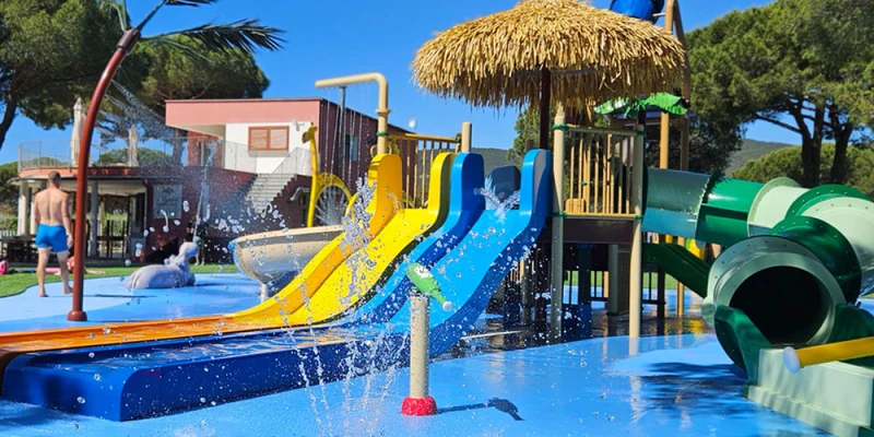 Colorful water slides splash into a shallow pool, surrounded by a water playground with trees and people nearby at Camping Lacona Pineta