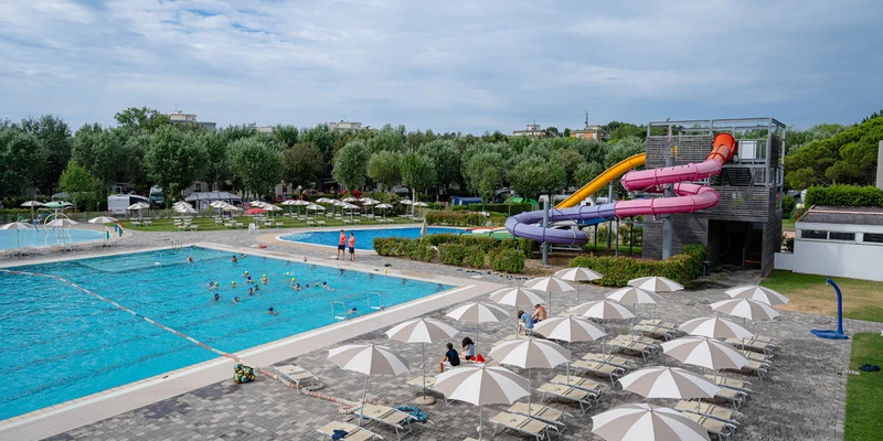Swimming pool with people playing; nearby water slides and sun loungers; surrounded by trees and umbrellas at Marina Julia Family Camping Village