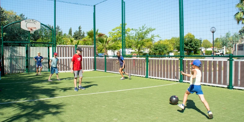 Children playing soccer on a fenced artificial turf court under a clear blue sky at La Prairie 