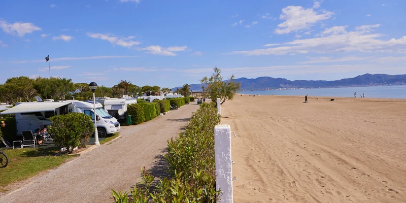 Campervans are parked by a sandy beach; people relax under awnings, with mountains visible in the distance at Nautic Almata
