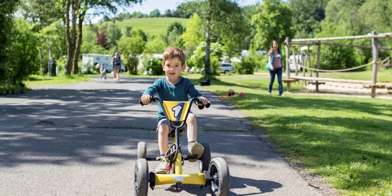 A child rides a tricycle on a paved path, surrounded by trees and greenery, with campers in the distance at Landal Wirfttal