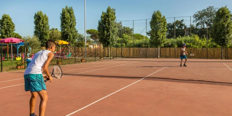 Two boys playing tennis on a clay court surrounded by greenery and colorful playground equipment at Villaggio Mare Si