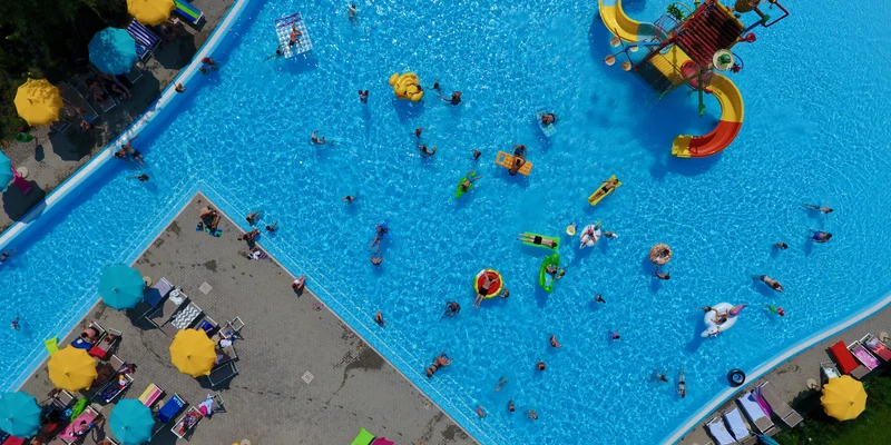 Aerial view of people swimming and playing in a large pool with colorful slides, surrounded by umbrellas and lounge chairs at Belvedere Village