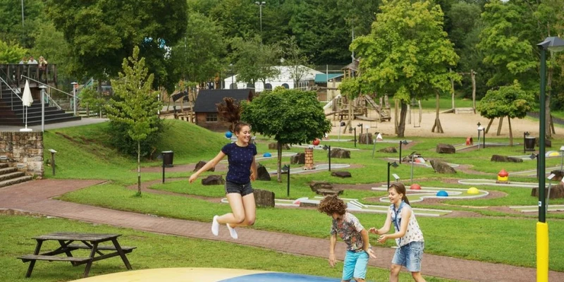 Three children bounce on an inflatable mat in a park playground with lush greenery and playground equipment at Landal Wirfttal