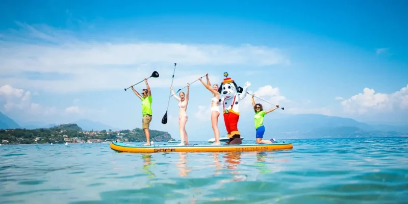 Five people and a costumed character paddleboard, raising paddles on a sunny, blue lake at Sivinos Camping Boutique