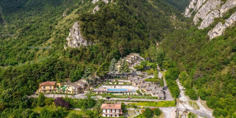 A resort nestled in a dense forested valley with mountains in the background, featuring multiple buildings and a central pool at Residence Oasi