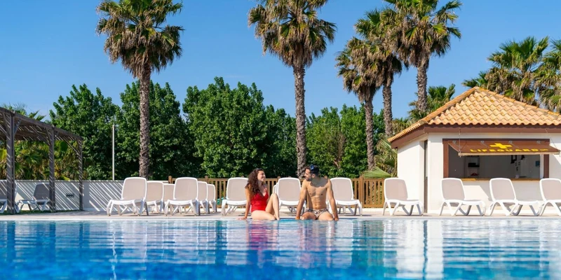 Two people sit by a clear swimming pool, surrounded by palm trees and lounge chairs, near a lifeguard station at Aloha