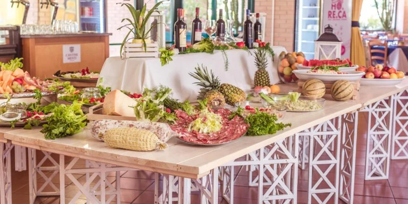 Food buffet arranged with various fruits, vegetables, and meats on decorated tables in a dining hall at Villaggio Mare Si