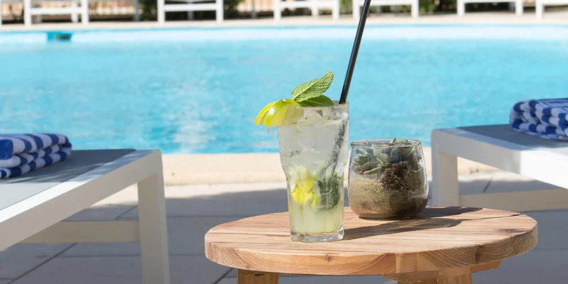 A cold drink sits on a wooden stool beside a swimming pool with sun loungers and umbrellas in the background at Les Bulles de Mer