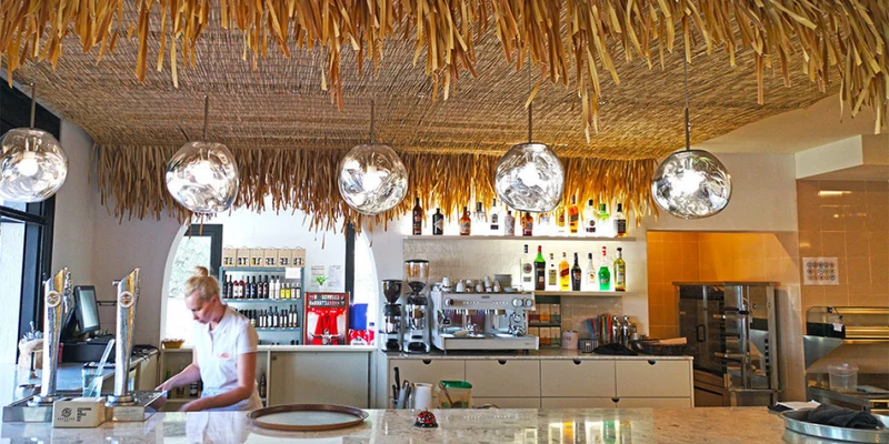 Counter with bartender preparing drinks under straw-roof decor and hanging lights, surrounded by shelves of bottles at Orangeraie