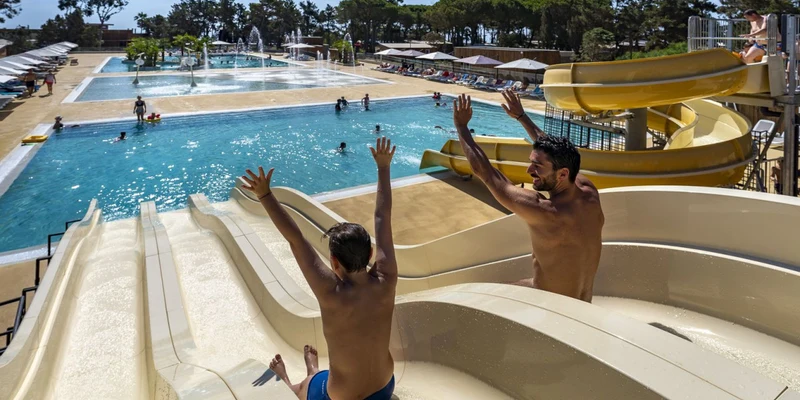 Two people slide down water slides into a large swimming pool area surrounded by lounge chairs and trees at Cap Sud