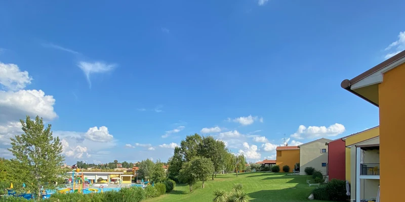 Colorful buildings line a lush green lawn, bordered by trees, under a clear blue sky at Belvedere Village