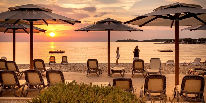 Beach chairs under umbrellas, two people photographing the sunset beside a calm sea with a distant island at Val Saline