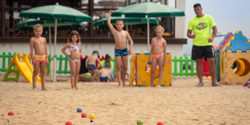 Children playing bocce in a sandy playground with colorful foam blocks and umbrellas, supervised by an adult at Punta Spin