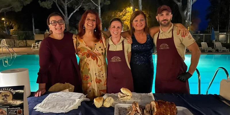 Five people smiling, standing behind a table with food, at night by a lit swimming pool at Punta Navaccia