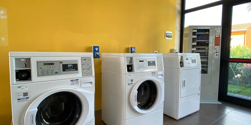 Washing machines and dryer stand idle against a yellow wall in a sunlit laundry room, near a vending machine at Belvedere Village