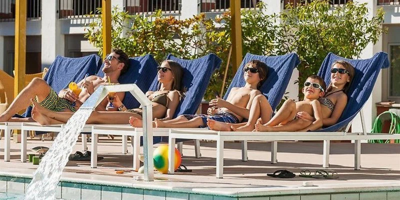 People relax on blue lounge chairs, sunbathing beside a pool with flowing water, surrounded by a building and trees at Holiday Park La Vela