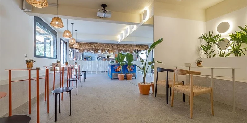 Seating area with tables and chairs, leading to a vibrant bar with plants and hanging lights at Orangeraie