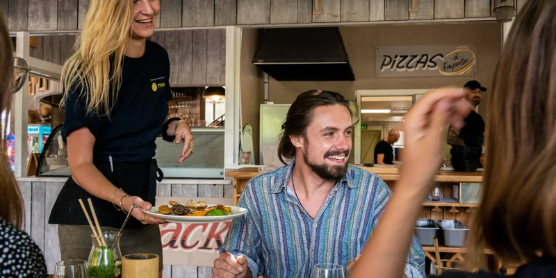 Waitress serves food to a smiling man at an indoor restaurant with rustic wooden decor at Le Millau Plage