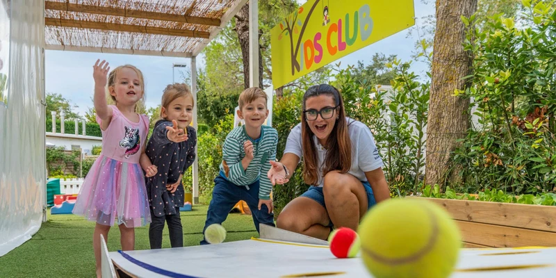 Children and an adult play ball toss game enthusiastically in an outdoor kids club area, surrounded by greenery at Europa Camping Village