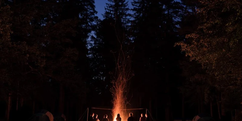 Group gathers around a campfire, sitting on logs, amid tents and tall trees at Forest camping Mozirje