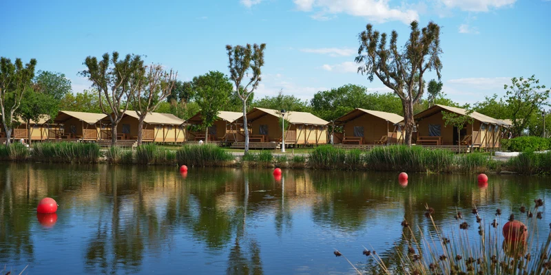 Tents stand along a lakeside, surrounded by trees and grass with red buoys floating on the water at Nautic Almata