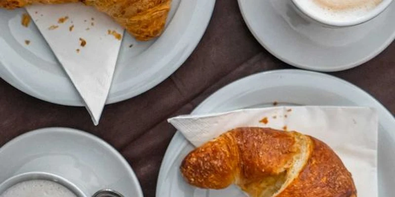 Croissants and cappuccinos on white plates atop a brown tablecloth, accompanied by spoons and napkins at Camping Mare Pineta