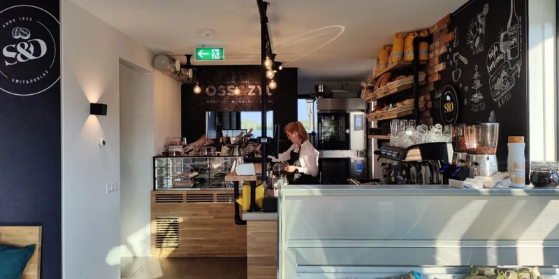 Barista preparing coffee behind a modern café counter with pastries, illuminated shelving, and a minimalist interior at Waterstaete Ossenzijl
