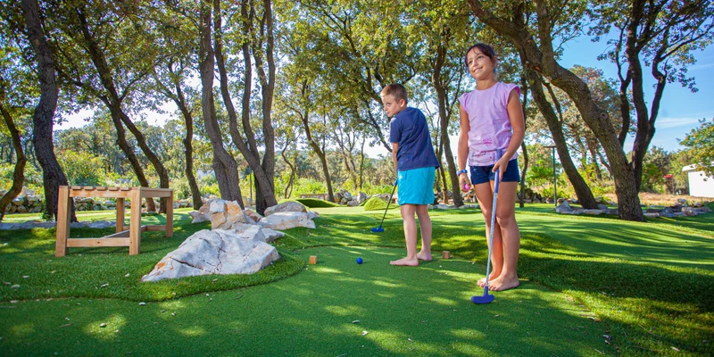 Children playing mini golf in a shaded, grassy area with scattered rocks and trees at Lopari