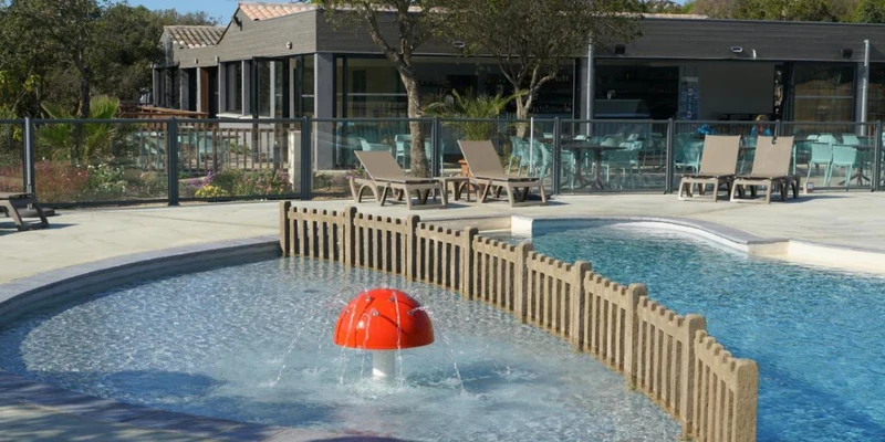 Red mushroom fountain spraying water in a shallow pool, surrounded by lounge chairs and trees at Camping Le Damier