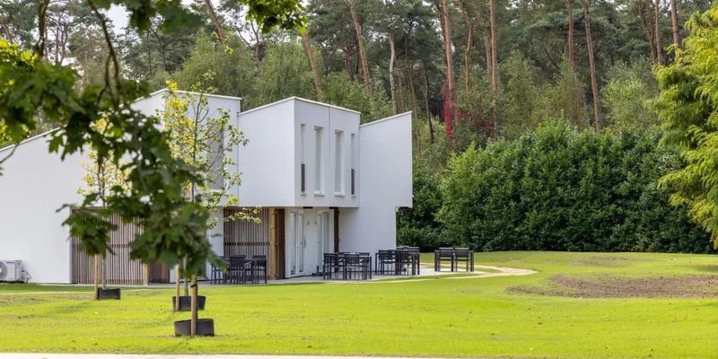 Modern white building surrounded by greenery; outdoor tables and chairs set up around it among trees and grass at Park Eksel
