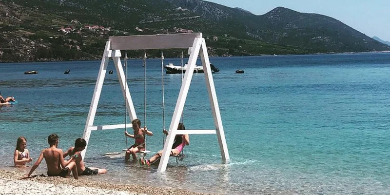 Children swing on a white swing set at the water's edge on a pebbled beach, with mountains and a boat in the background at Nevio