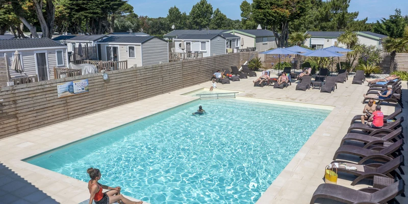 A person lounges poolside as others swim or sunbathe, surrounded by mobile homes and trees at Les Dunes 