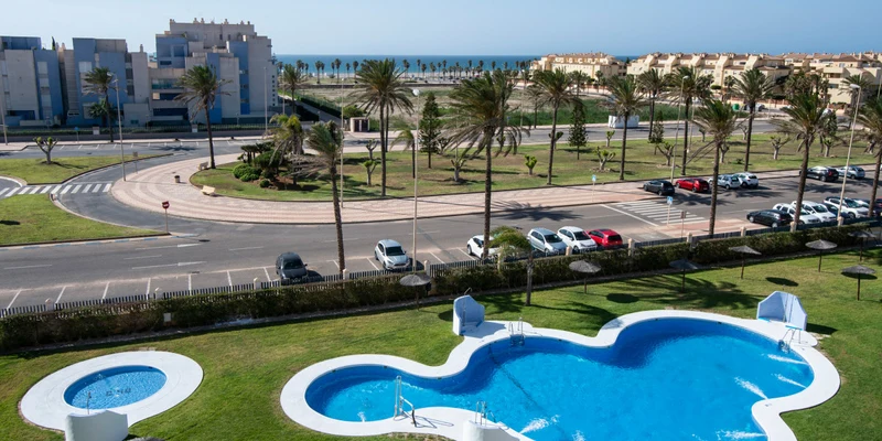 A curvy swimming pool sits amid grassy areas, surrounded by palm trees near a road and residential buildings at Alméria Roquetas de Mar