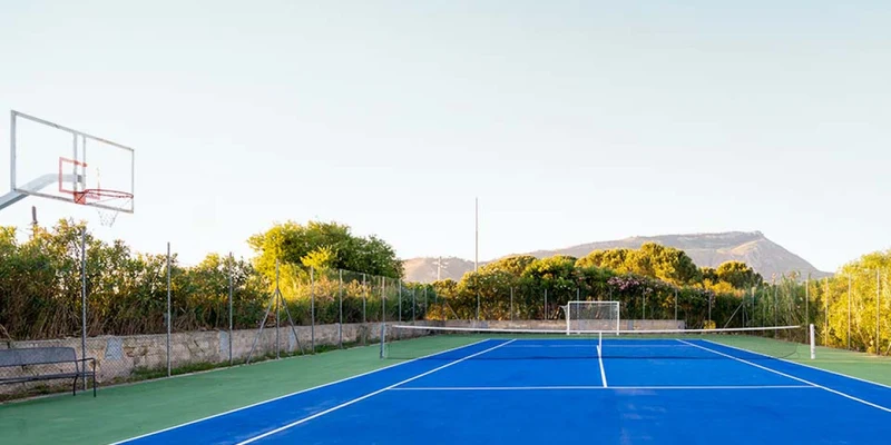 A blue tennis court with basketball hoop, flanked by greenery and mountains in the background at Duca di Castelmonte