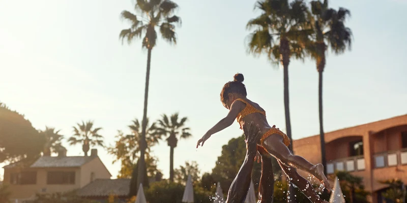 Person lifts child in swimming pool with splashing water, surrounded by palm trees and buildings at Les Rives de Cannes Mandelieu