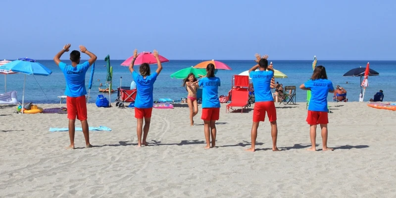 People in blue shirts and red shorts doing exercises on a sandy beach with colorful umbrellas at Riva di Ugento