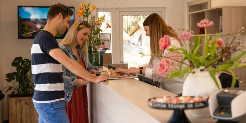 A couple registers at a reception desk, engaging with a receptionist in a flower-adorned office at Europarcs Hoge Kempen