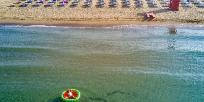 Person floats on an inflatable ring in the sea near a sandy beach with rows of blue umbrellas at Le Marze