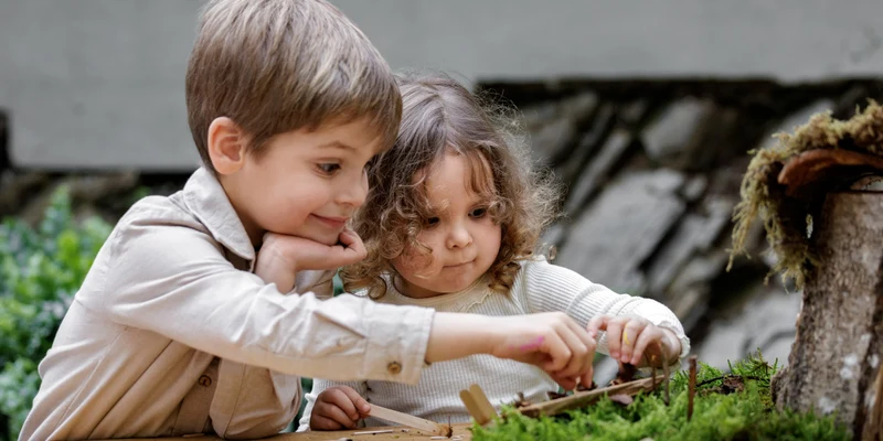 Children constructing crafts at a wooden table outdoors, surrounded by greenery and a stone wall at Hillview Resort Grandvoir