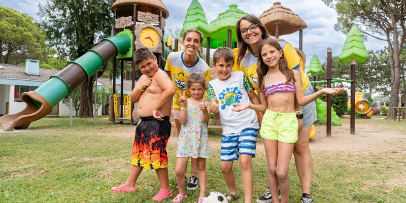 Group of smiling children and adults posing together in front of a colorful playground at Marina Julia Family Camping Village